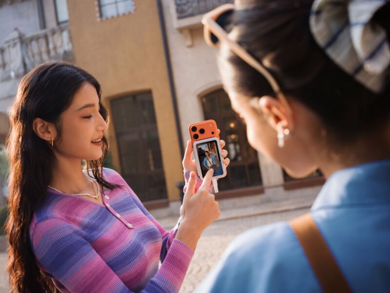 Two young women outdoors, one showing the other a photo on her smartphone. The woman in the foreground is slightly out of focus, while the woman holding the phone smiles and points at the screen.