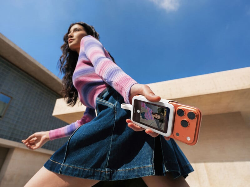 A woman in a purple and pink striped sweater and denim skirt holds an instant camera and a printed photo, walking outdoors with a building and blue sky in the background.