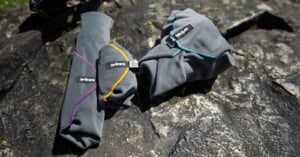 Three rolled-up gray outdoor garments with colored trim (purple, yellow, and blue) and "AURK" labels are placed on a rocky surface in bright sunlight.