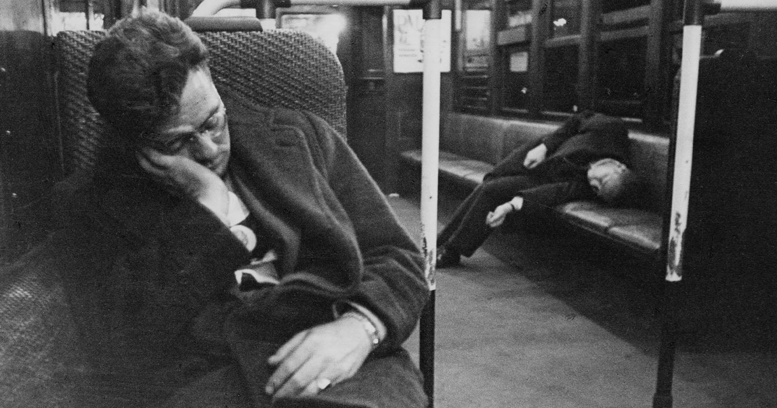 Two people sleep on an empty subway train; one sits slumped against a seat in the foreground, while the other lies across a bench in the background. The scene appears quiet and dimly lit.