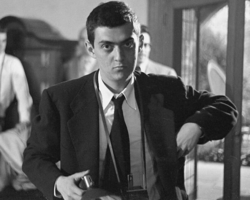 Young man in a suit and tie holds a camera, looking directly at the viewer with a serious expression. The background shows other people in a softly focused, indoor setting with natural light.