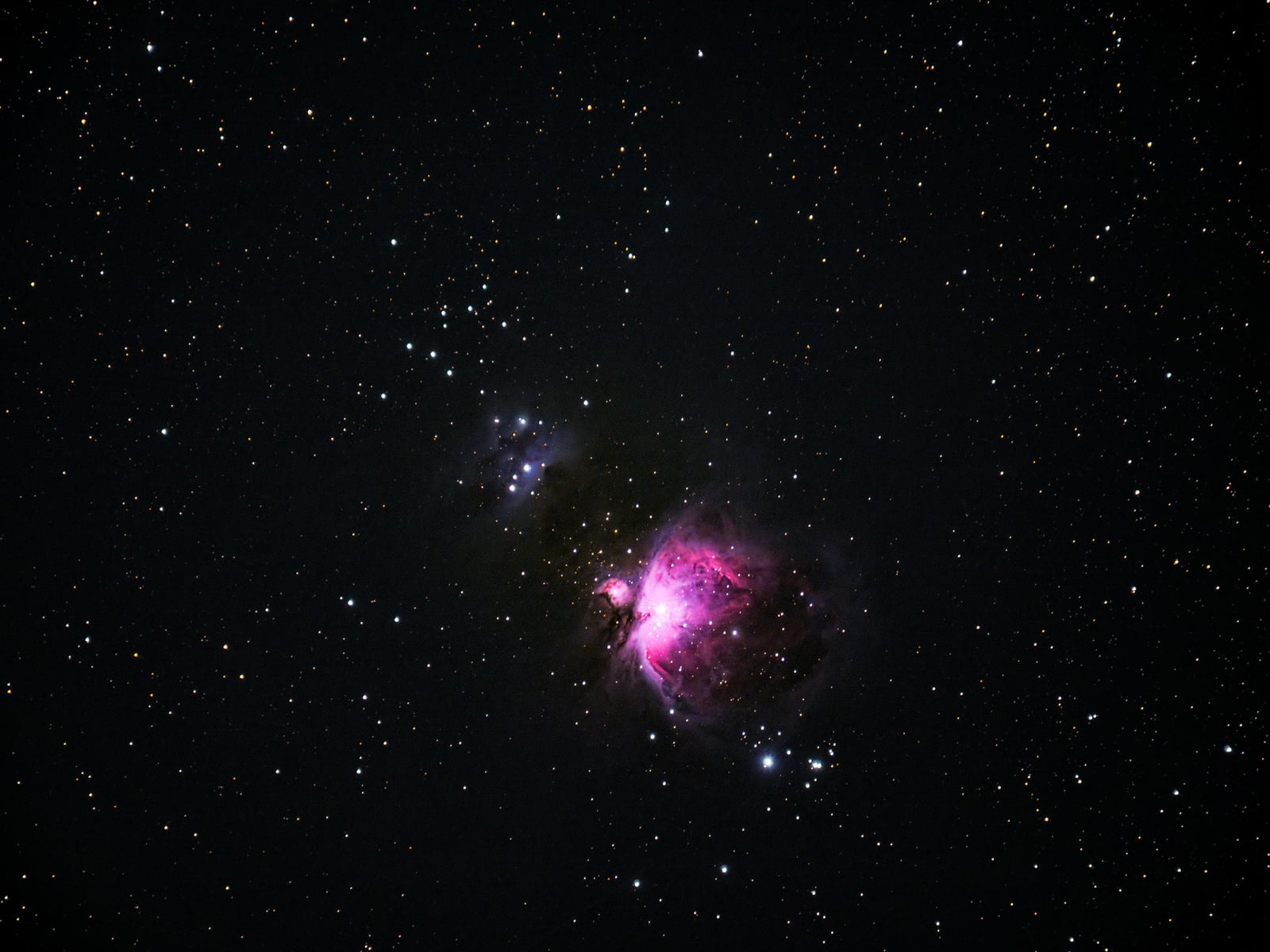 A deep space photo showing a star-filled sky with the bright, pinkish-purple glow of the Orion Nebula surrounded by scattered stars and dark space.