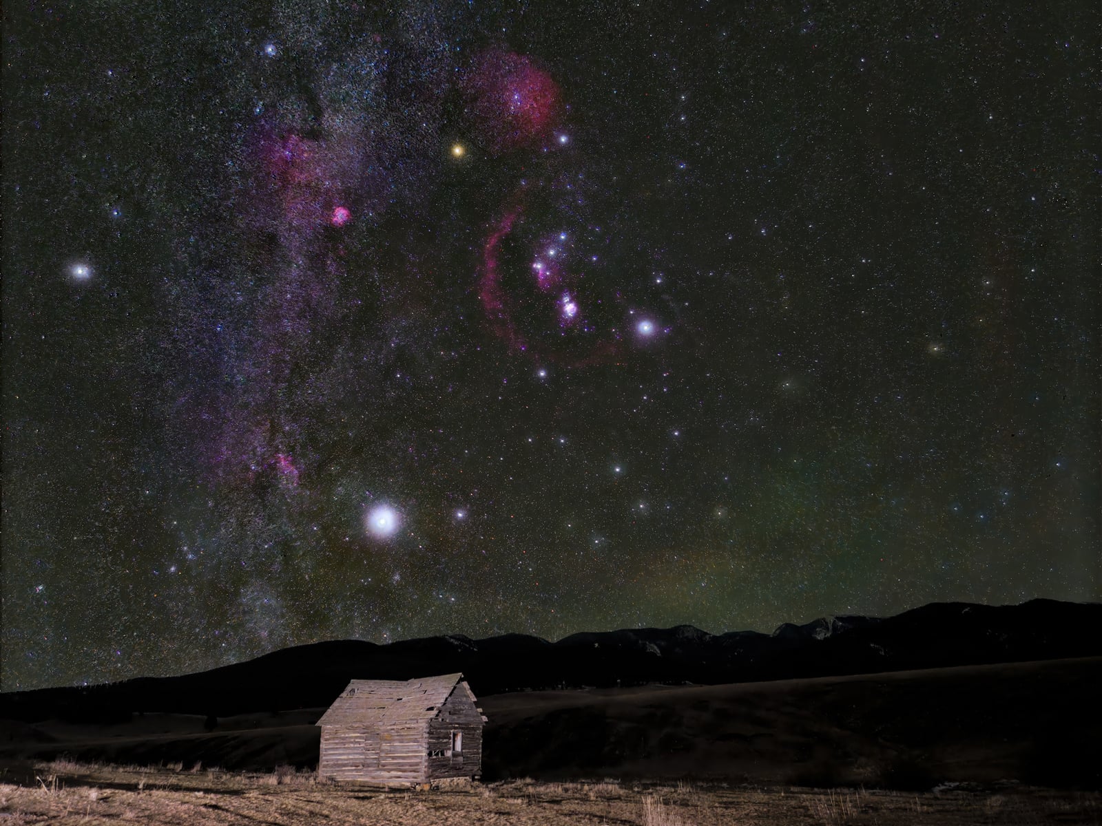 A small, old wooden cabin stands in a field under a night sky filled with bright stars and colorful nebulas, with mountains silhouetted in the background.