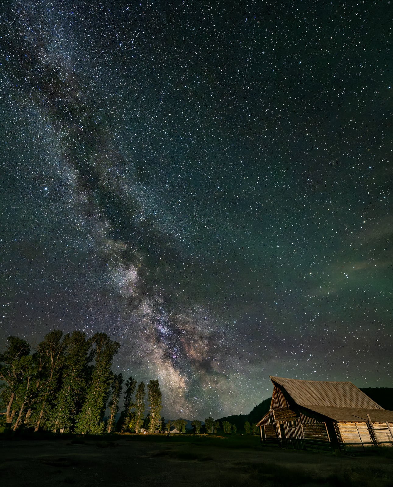 A rustic wooden cabin sits beneath a night sky filled with bright stars and the glowing band of the Milky Way, with tall trees silhouetted on the left.