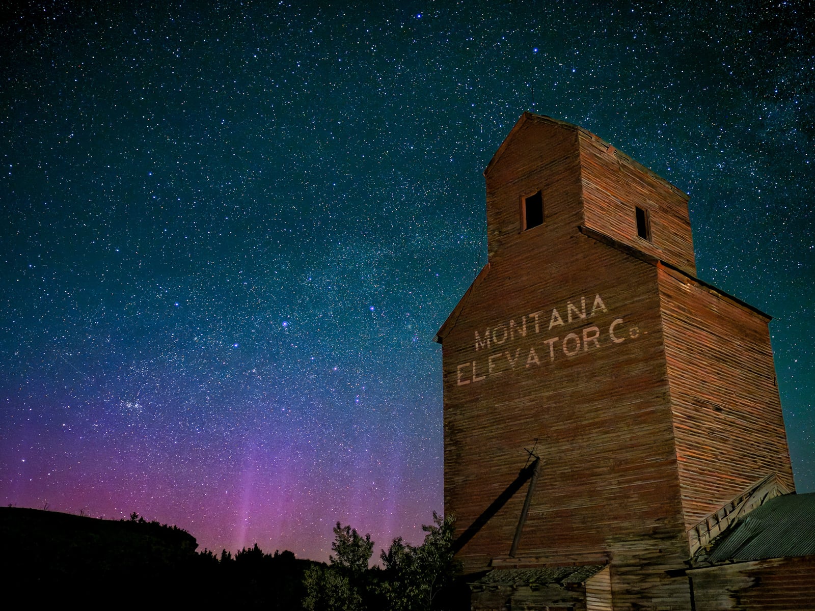 An old wooden grain elevator labeled "Montana Elevator Co." stands under a clear, star-filled night sky with a faint purple aurora near the horizon.