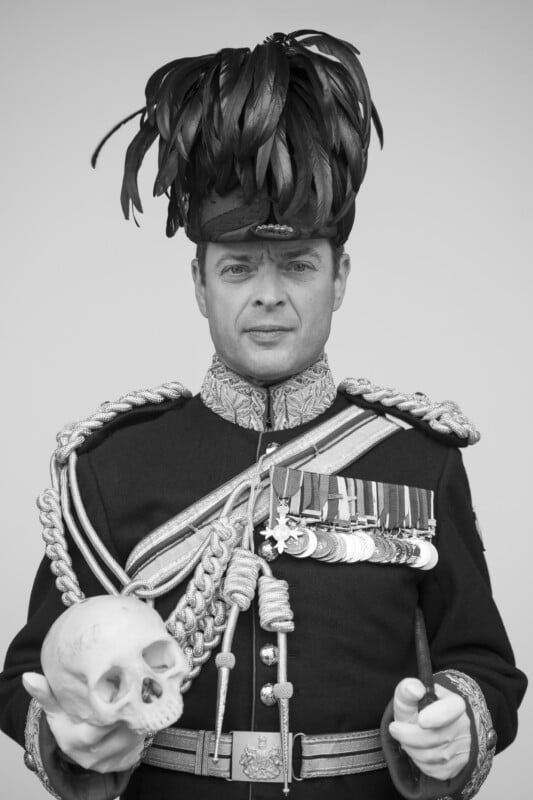 A man in a decorated military uniform and feathered hat holds a human skull in one hand and a baton in the other, with several medals displayed on his chest. Black and white photo.