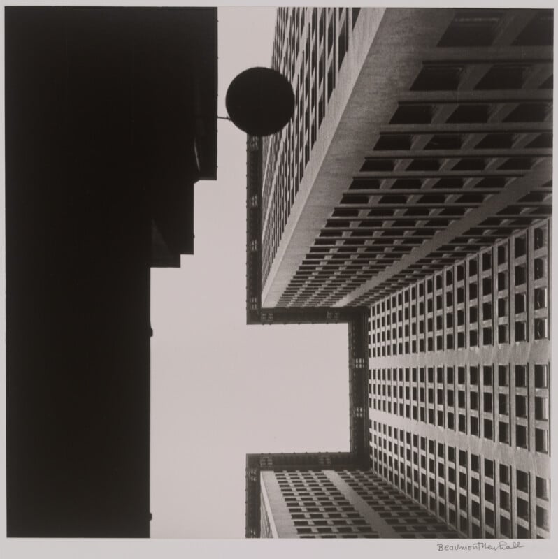 Black and white photo looking up at tall, geometric buildings with many windows, forming sharp angles against the sky; a circular object is suspended between the buildings.