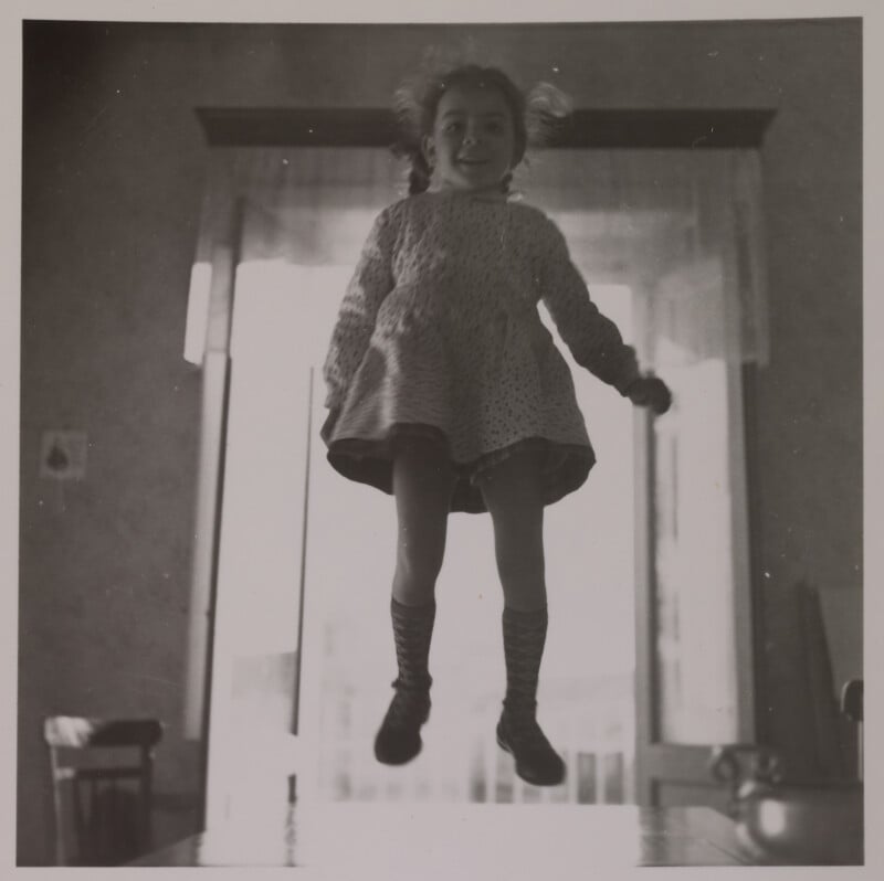 A young girl with braided hair and a knitted sweater is caught mid-jump indoors, smiling, with light streaming through a large window behind her.