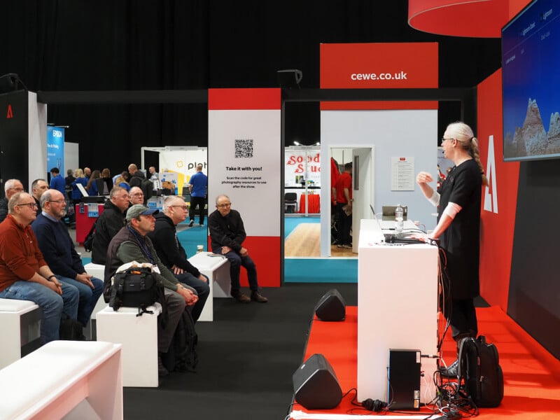 A speaker presents at a tech event to an audience seated on white benches. The audience is attentive, and a screen with a mountain image is visible. Booths and banners, including "cewe.co.uk," are in the background.
