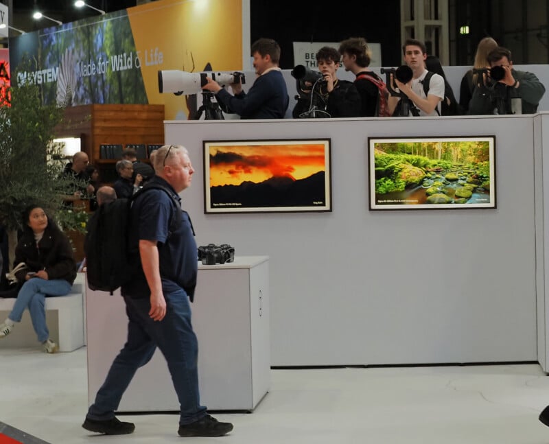 A man with a backpack walks past a display of landscape photographs, while several people with large cameras stand behind the display taking pictures; other people are seated nearby.