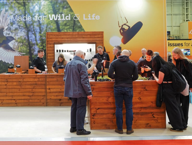 People gather around a wooden counter of the OM System stand at a photography expo, examining cameras and lenses. A large backdrop features nature imagery and the words "Made for Wild & Life." Staff assist visitors.