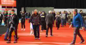 A crowd of people walks and stands on a red carpet at an indoor event, with booths and a large "my cewe photobook" banner visible in the background. Some attendees carry bags and a photographer is taking pictures.