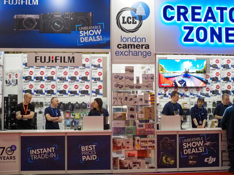 A busy camera store booth at an event features staff behind counters, Fujifilm and London Camera Exchange signs, cameras and accessories on display, price tags, and digital screens showing promotional deals.