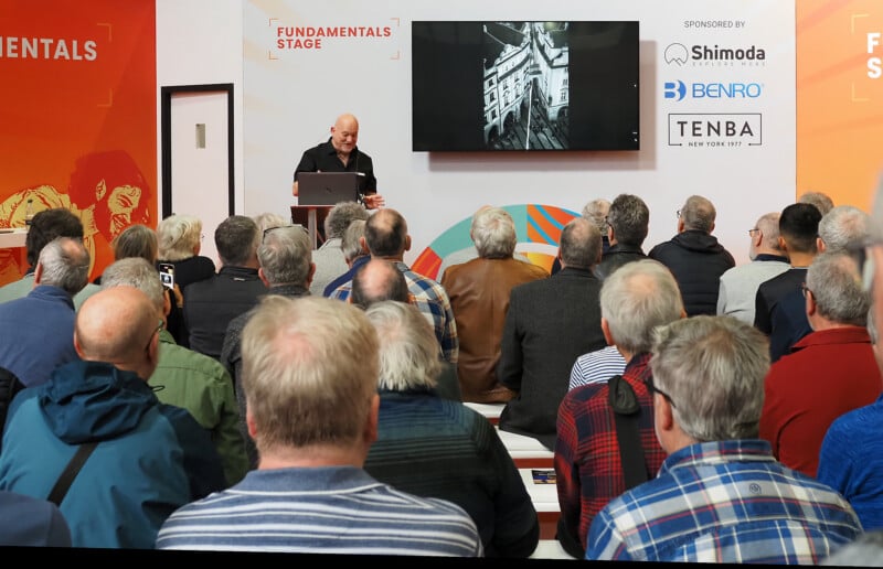 A speaker presents to an audience seated indoors at a photography event, showing a black-and-white photo on a screen. Event banners and sponsor logos are visible on the walls.