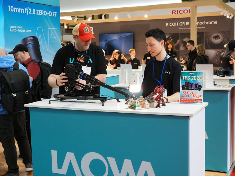 Two men at a LAOWA booth display a camera and lens setup, with camera equipment and small figures on the counter; people and promotional banners are visible in the background at a busy event.