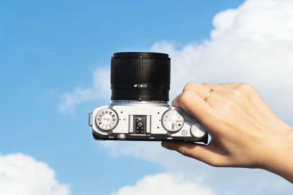 A hand holding a silver and black camera against a blue sky with white clouds; the person is wearing a gold ring on their ring finger.