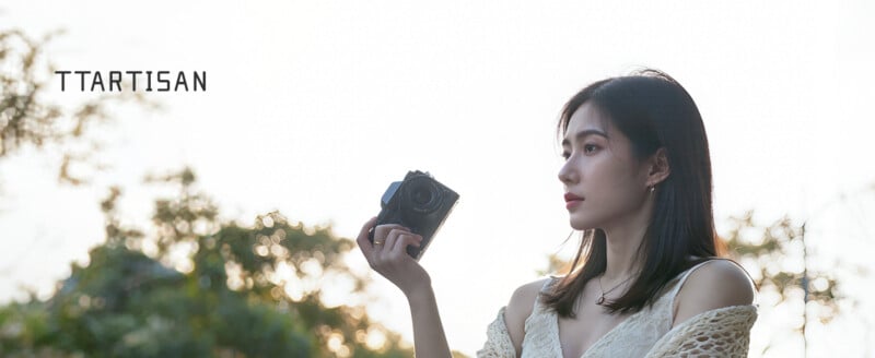 A woman in a light dress holds a camera and looks into the distance outdoors, with trees in the background and the word "TTARTISAN" displayed on the left.