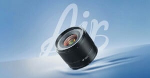 A camera lens labeled "AIR 12mm F2" is displayed against a smooth blue background with the word "Air" written in large, white script behind it. The lens reflects light and appears to be floating.