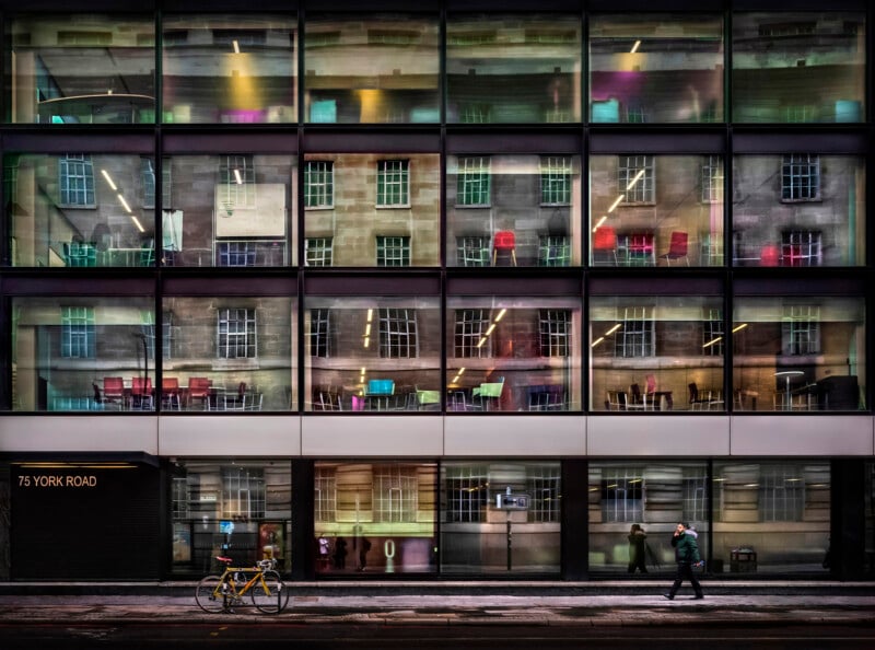 A modern glass building shows reflections of an older building and colorful chairs inside. Pedestrians walk by on the sidewalk, and a yellow bicycle is parked near a sign reading "75 York Road.