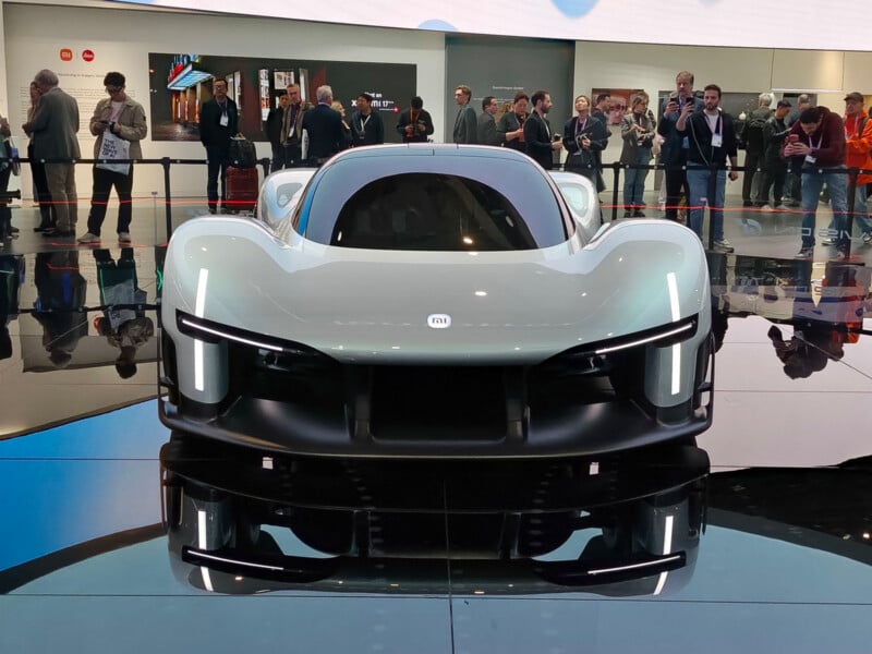 A futuristic white sports car is displayed on a shiny platform at an auto show, with people standing and observing in the background. The car has sleek curves, LED lights, and a low aerodynamic design.