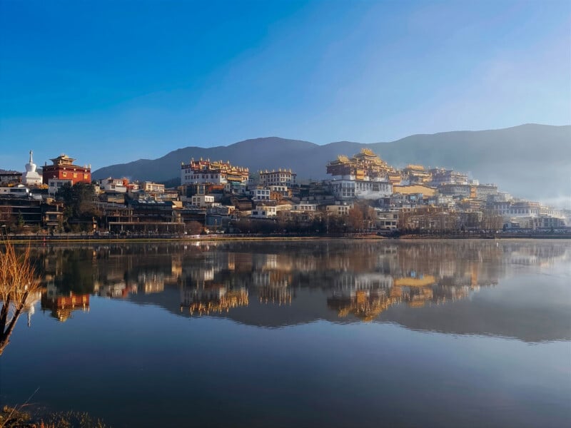 A hillside town with traditional Tibetan-style buildings and gold rooftops is reflected in a calm lake, with mountains and a clear blue sky in the background.