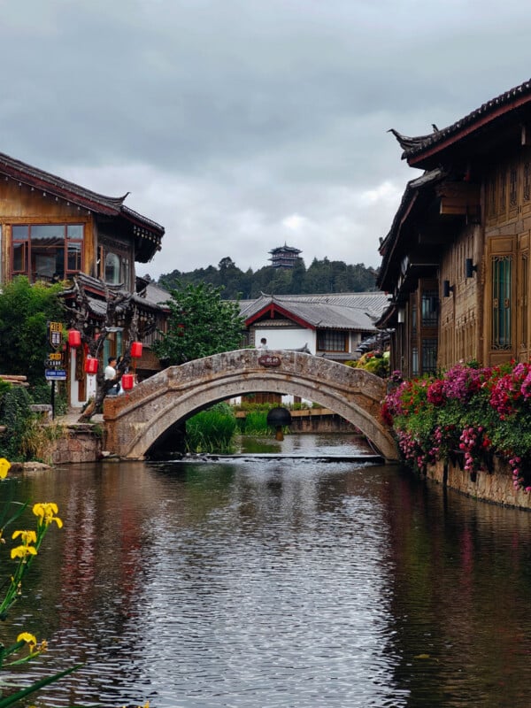 A traditional stone arch bridge spans a calm canal lined with wooden buildings, red lanterns, and blooming flowers; a distant pavilion sits atop a hill under a cloudy sky.