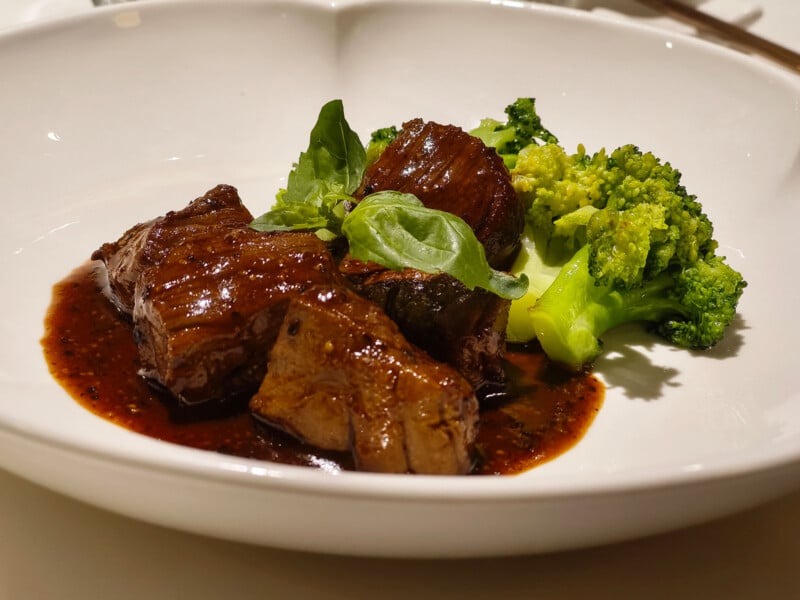 A white plate with braised beef in a rich brown sauce, accompanied by steamed broccoli and fresh green garnish.