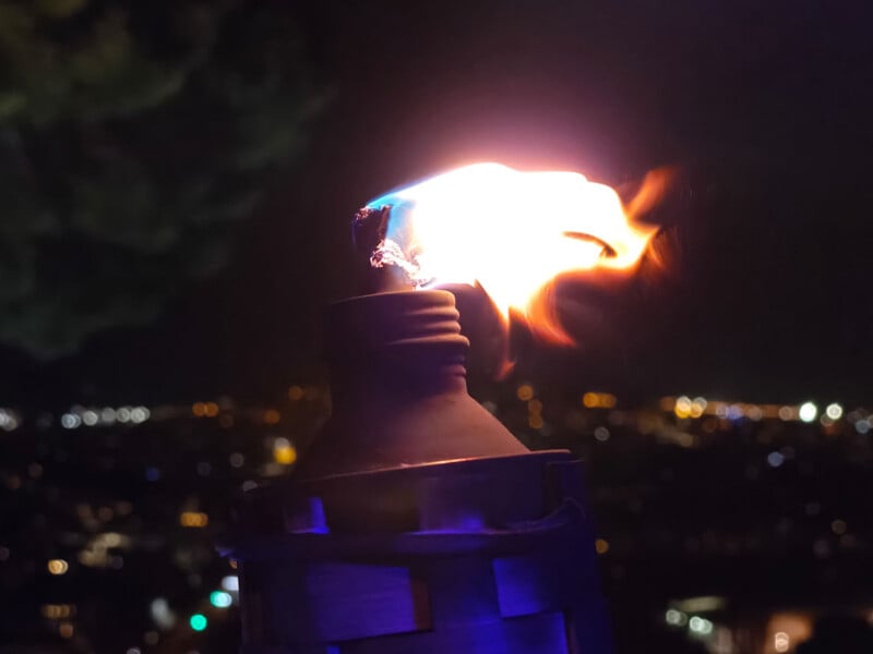 A lit torch burns brightly against a blurred cityscape at night, with orange flames and a glowing wick. The background shows out-of-focus city lights and a dark sky.