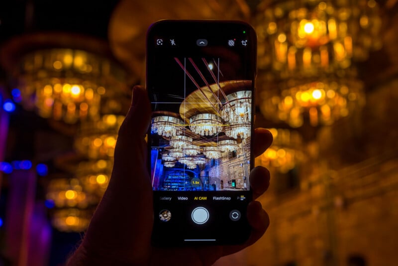 A hand holds a smartphone, capturing an image of ornate, glowing chandeliers against a warmly lit, blurred background with golden lights. The phone screen displays the chandelier scene in focus.