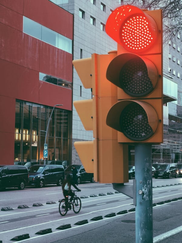 A traffic light shows red while cars wait at an intersection. A cyclist rides past on a bike lane in front of a modern building with large windows.