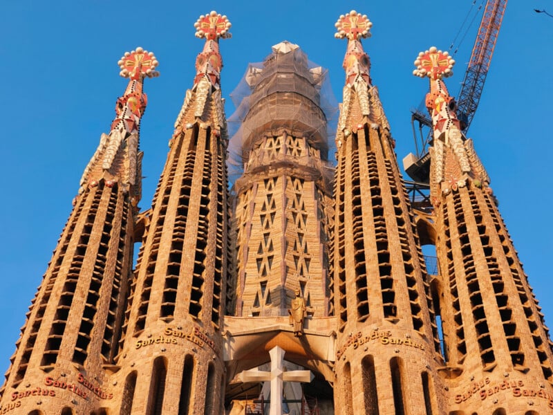 Four ornate towers of the Sagrada Família basilica in Barcelona rise against a clear blue sky, showcasing detailed stonework and colorful floral decorations near the tops.