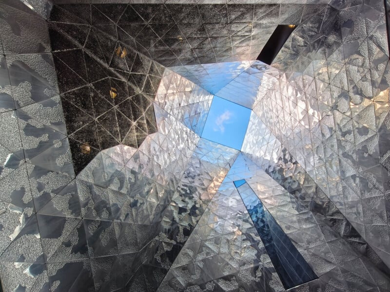 A view looking up through a geometric, reflective metal ceiling with angular shapes, revealing a patch of blue sky and clouds in the center. The surfaces mirror light and create abstract patterns.