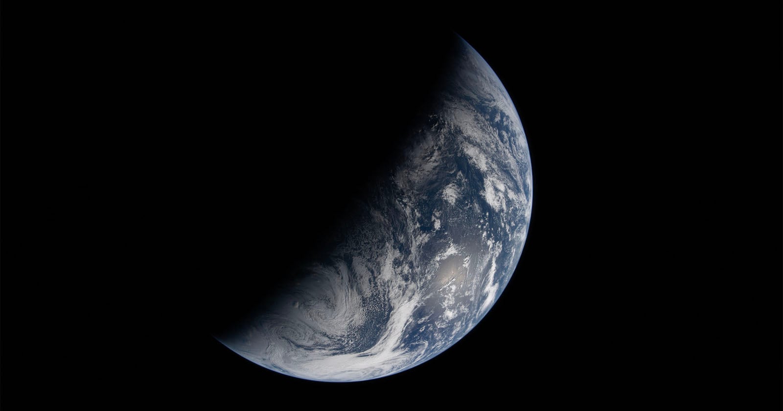 A crescent view of Earth from space, with one side illuminated by sunlight and the other in shadow, showing blue oceans, white clouds, and portions of continents against a black background.