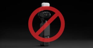 A small black camera with an attached white accessory is shown against a dark background. A large red prohibition symbol overlays the camera, indicating it is not allowed or restricted.