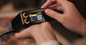 Close-up of hands adjusting settings on a compact audio recording device with a small screen displaying menu options and two orange control knobs.