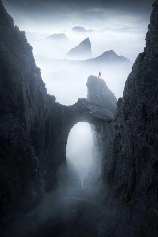 A person in a red jacket stands on a rocky archway between steep cliffs, surrounded by mist and distant jagged mountains under a cloudy sky.