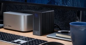 A compact desktop computer, an external black device with ventilation holes, a keyboard, a notebook, a pen, and a blue cup are arranged on a wooden desk in front of a monitor.