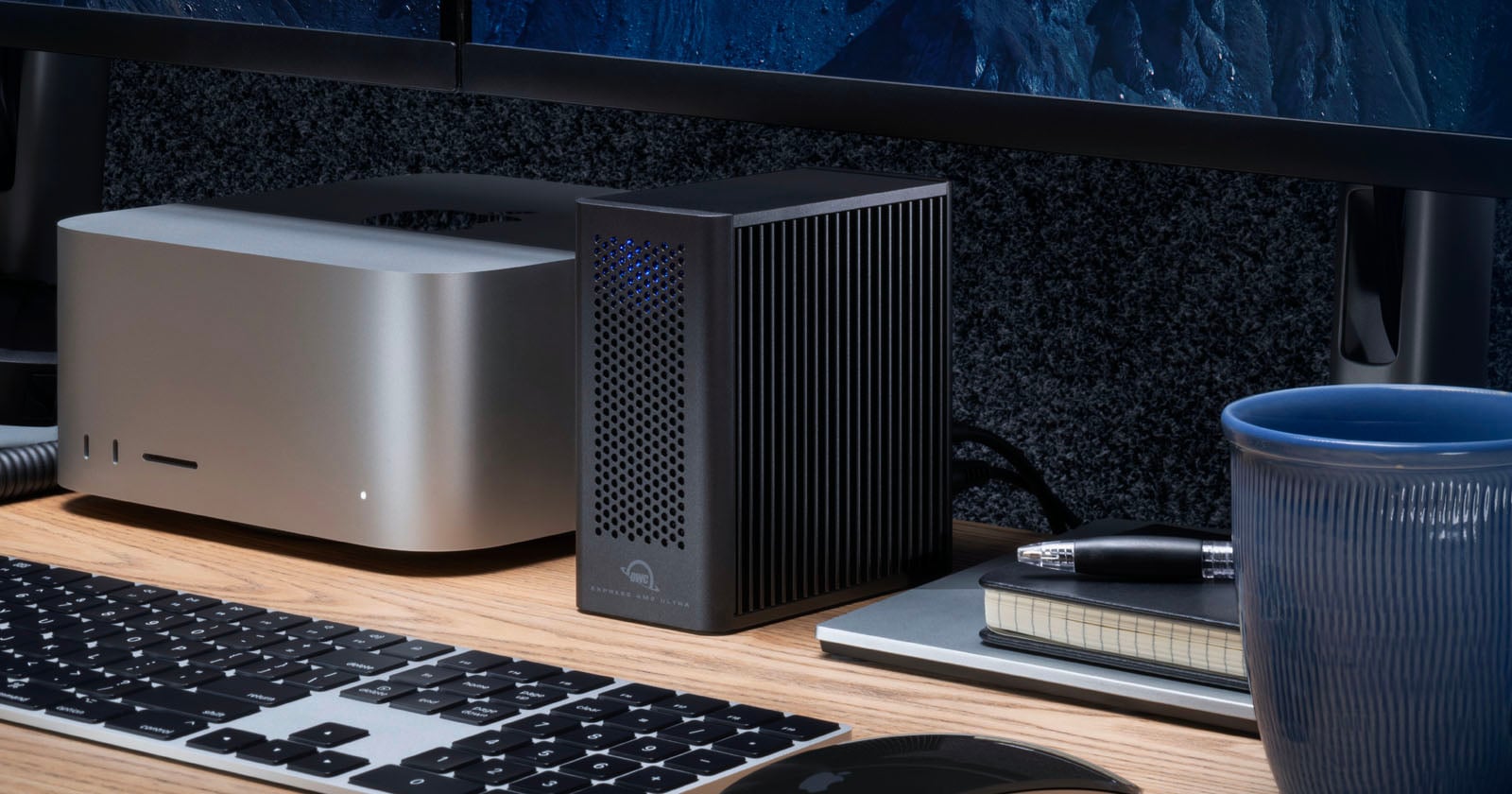 A compact desktop computer, an external black device with ventilation holes, a keyboard, a notebook, a pen, and a blue cup are arranged on a wooden desk in front of a monitor.