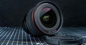 A Rokinon FE 14-24mm F2.8 camera lens is placed on a grid-patterned surface, with its lens cap lying beside it. The background appears out of focus, emphasizing the lens.