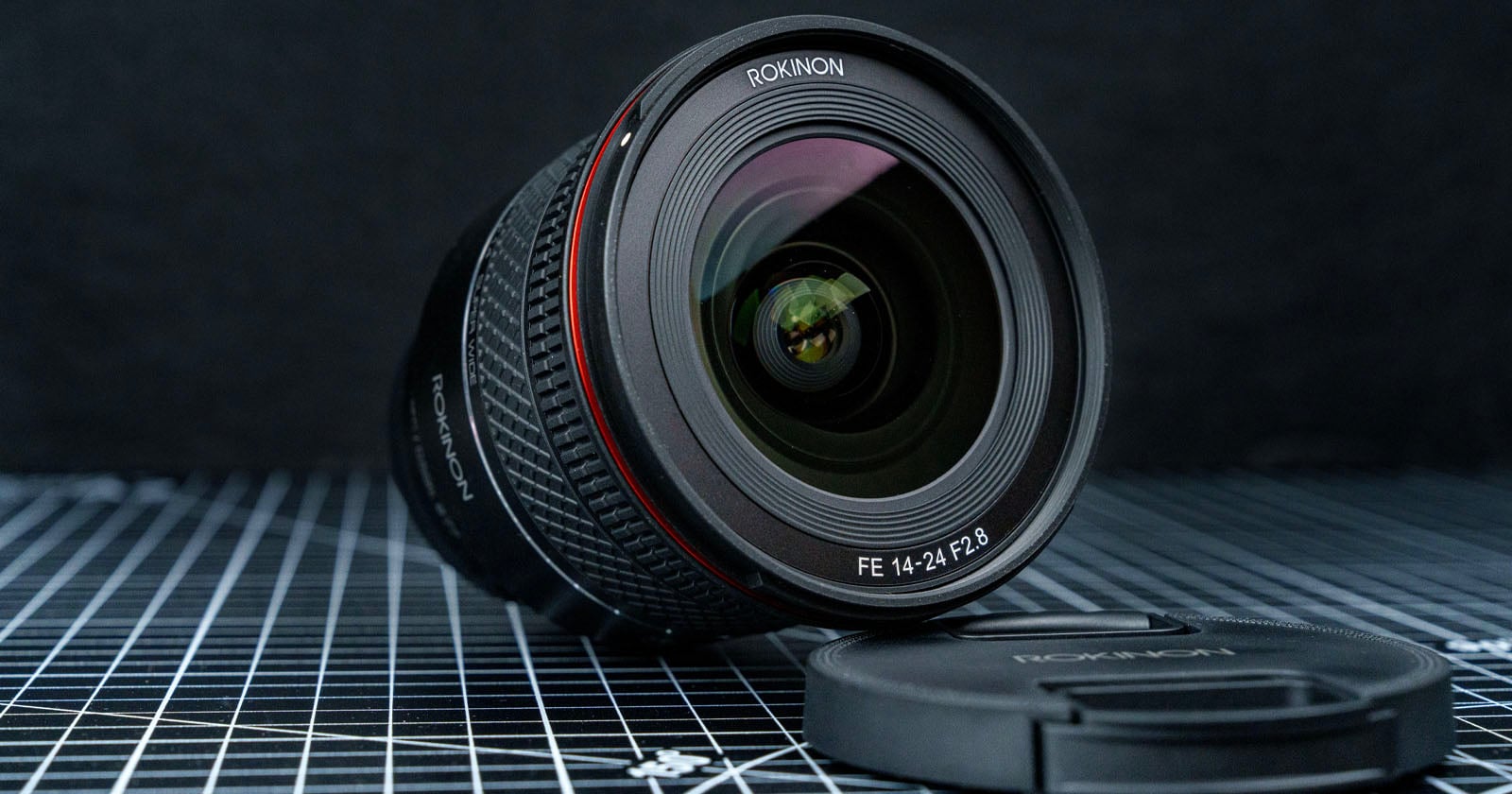 A Rokinon FE 14-24mm F2.8 camera lens is placed on a grid-patterned surface, with its lens cap lying beside it. The background appears out of focus, emphasizing the lens.