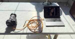 A camera connected to a laptop with an orange cable sits on a marble table. The laptop screen shows a blurred image of a person, and sunlight casts shadows across the table.