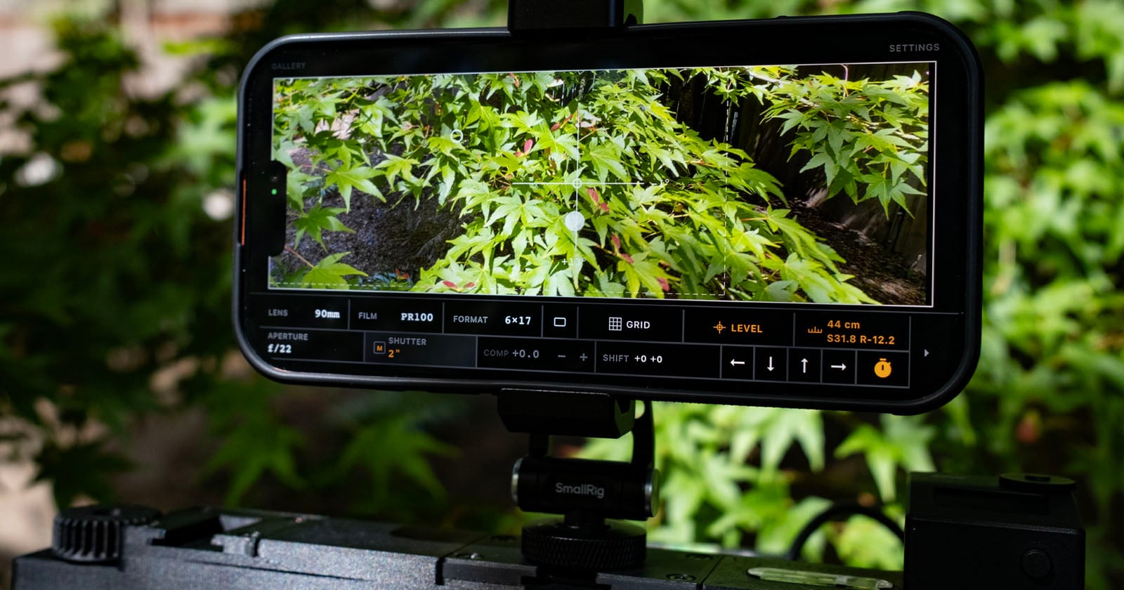 A smartphone mounted on a camera rig displays a live view of green maple leaves, with detailed camera settings visible on the screen. The background shows matching green foliage.