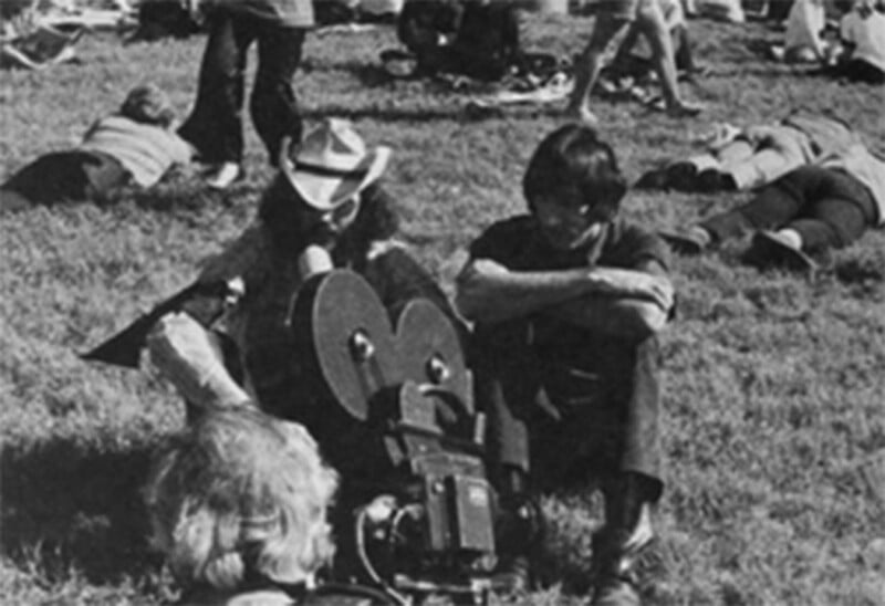Black-and-white photo of people sitting and lying on grass, with two people near a vintage film camera in the foreground. Others are scattered and relaxed in the background.
