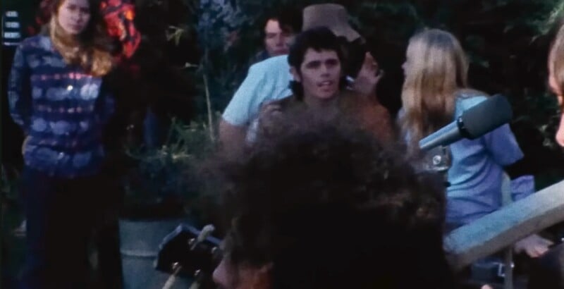 A group of people outdoors, some talking and others listening, with plants and equipment visible in the background. One person in the center looks towards the camera.