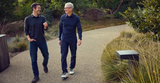 Two men walk and talk on a path surrounded by greenery. Both are wearing dark shirts and jeans, smiling as they enjoy the outdoor setting. Shrubs and tall grasses line the walkway.