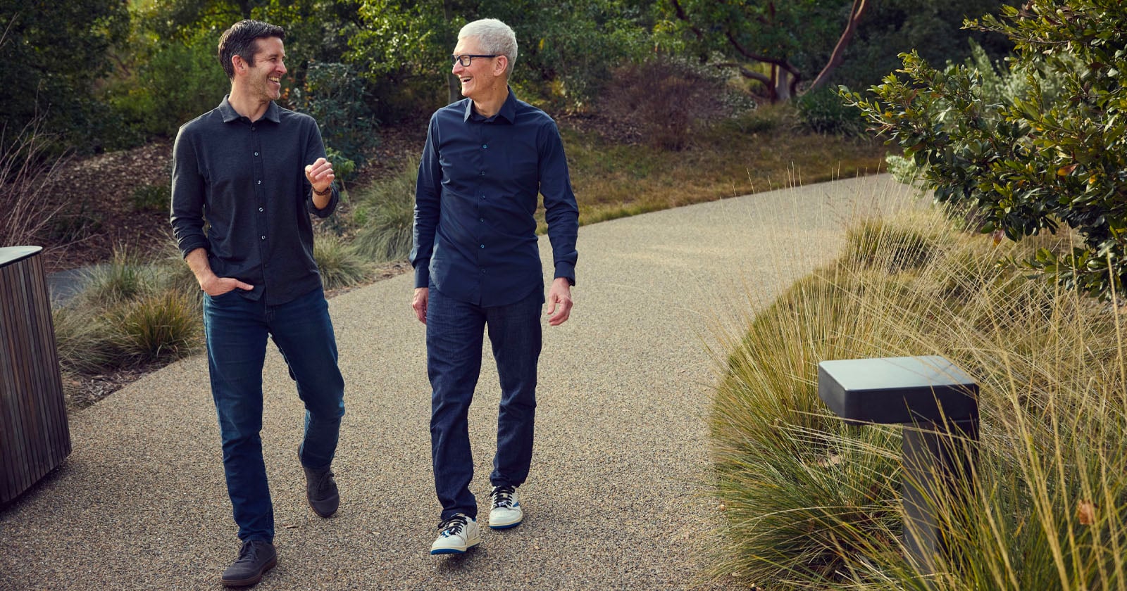 Two men walk and talk on a path surrounded by greenery. Both are wearing dark shirts and jeans, smiling as they enjoy the outdoor setting. Shrubs and tall grasses line the walkway.