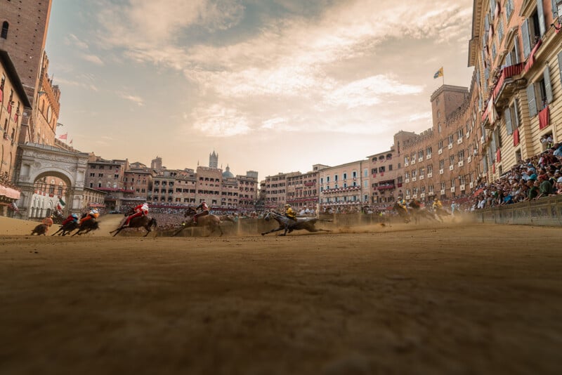 Horse race in a historic city square with riders in colorful outfits, kicking up dust as crowds watch from the surrounding medieval buildings under a dramatic evening sky.