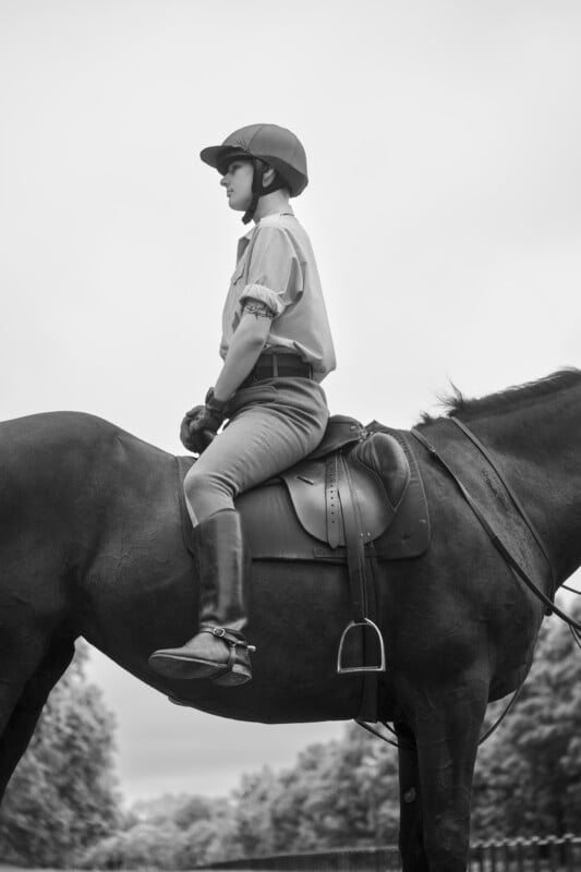 A person wearing a helmet and riding clothes sits upright on a saddled horse outdoors, looking ahead. The photo is in black and white, showing trees and a cloudy sky in the background.