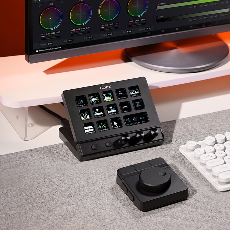 A digital control panel with colorful screens and knobs sits on a desk beside a white keyboard and a monitor, with a second black control device nearby. The setup is clean and modern.