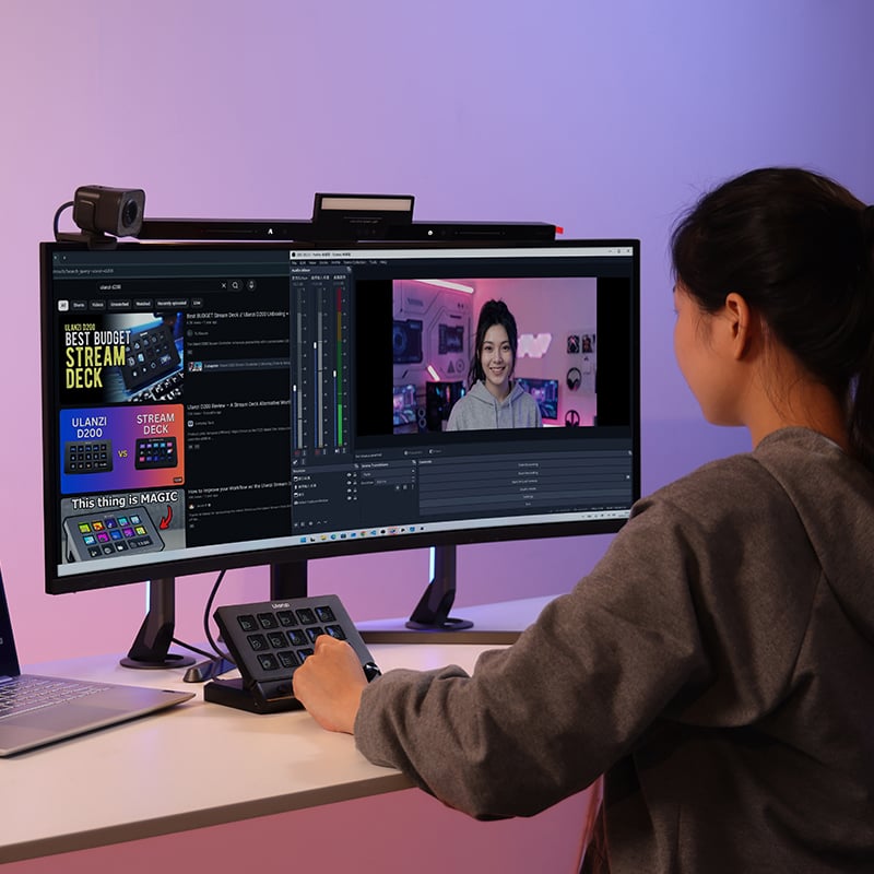 A person sits at a desk using a computer with a video streaming setup. The monitor shows streaming software with a person on a video call. There is a webcam, control pad, and colorful lights in the background.